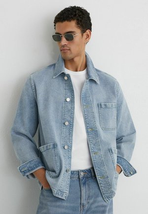 Muž v svetlomodrej rifľovej bunde a rifliach, v bielom tričku, s rukami v náprsných vreckách bundy, pozerajúci sa doľava, na pozadí jednoduchej steny.