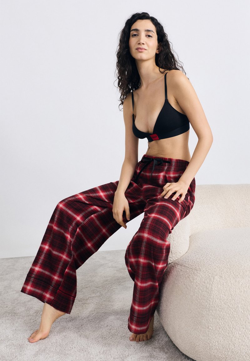 Μαύρο σουτιέν τρίγωνο με κόκκινη ετικέτα· κόκκινο καρό παντελόνι με φαρδιά πόδια· απα�λό ύφασμα· άνετη εφαρμογή· καθιστή στάση, ξυπόλητη πάνω σε υφή επιφάνειας.