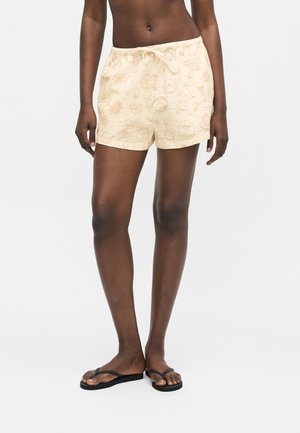 Personne à la peau foncée portant un short beige clair avec un motif circulaire subtil et des tongs noires, debout devant un fond uni.