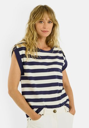 Femme aux longs cheveux blonds portant un haut sans manches rayé bleu marine et blanc et un pantalon blanc, les mains dans les poches.