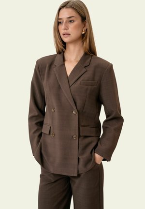 Nainen, joka käyttää ruskeaa kaksirivistä bleiseriä ja siihen sopivia housuja, kädet taskuissa, katsoo hieman sivulle.