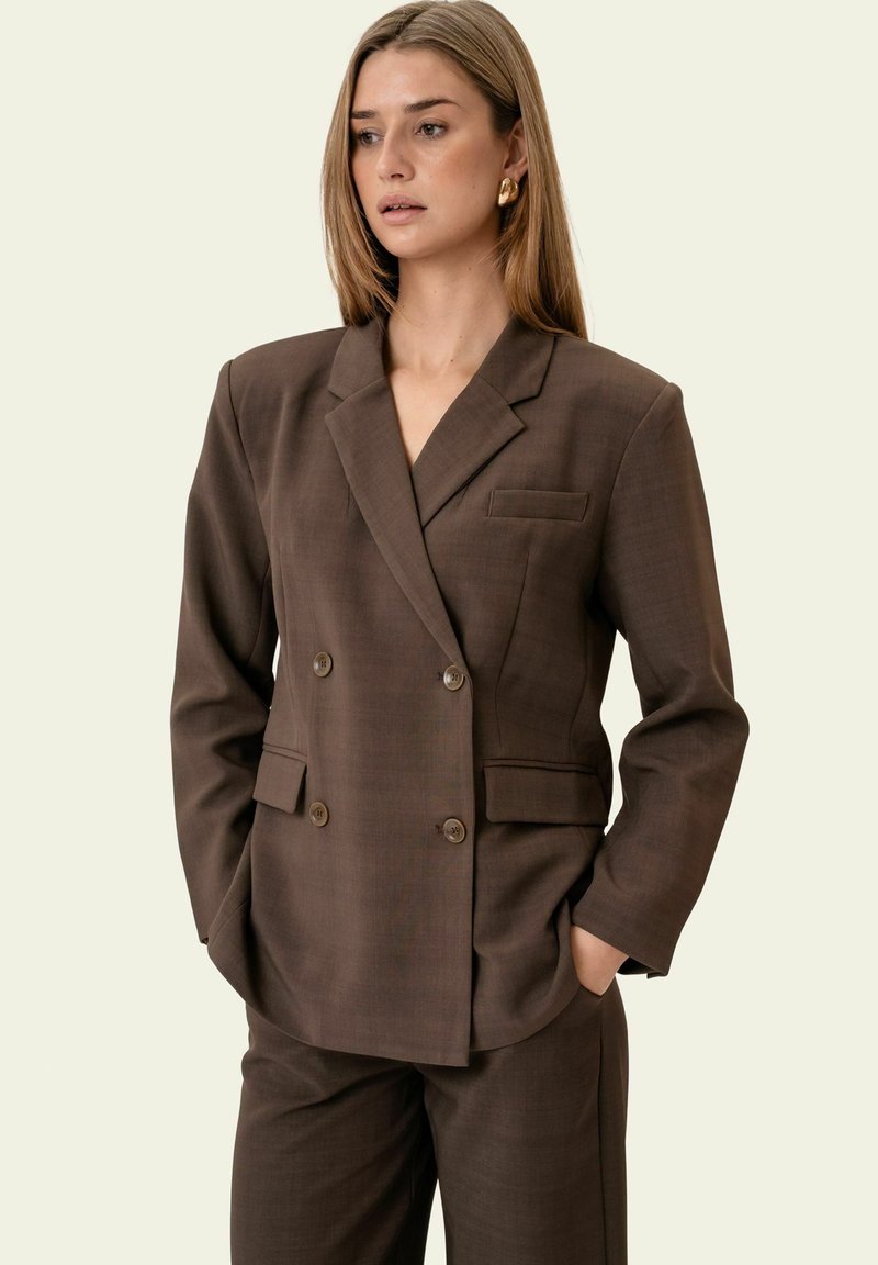 Mujer con blazer cruzado marrón y pantalones a juego, con las manos en los bolsillos, mirando ligeramente hacia un lado.