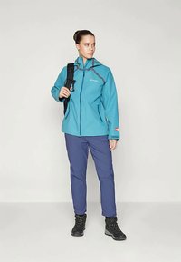 Νεαρή γυναίκα με τζάκετ σε τιρκουάζ χρώμα, μπλε παντελόνι και μαύρες ορειβατικές μπότες, κρατώντας μια μαύρη σακίδιο πλάτης, που στέκεται μπροστά σε έναν απλό φωτεινό φόντο.