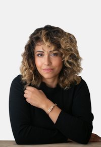 Maglia nera a maniche lunghe, capelli ricci, mostra un bracciale dorato al polso. Carnagione media, trucco naturale e sfondo neutro.