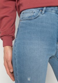Vaqueros de denim azul claro con un corte ajustado, que presentan un ligero desgastado en la parte delantera y un diseño de cintura alta. Detalles visibles de bolsillos y costuras.