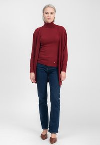 Cardigan bordeaux sopra un dolcevita coordinato, abbinato a jeans blu scuro. Il cardigan ha un frontale aperto, una trama liscia e maniche lunghe.