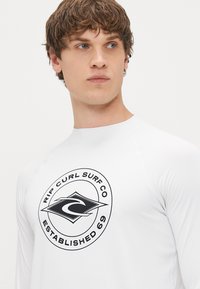 Λευκή μακρυμάνικη μπλούζα rash guard με κυκλικό μαύρο λογότυπο που απεικονίζει ένα κύμα και κείμενο "RIP CURL SURF CO ΙΔΡΥΘΗΚΕ 69."