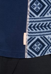 Granatowy polar z wzorzystym jasnoniebieskim panelem o geometrycznych wzorach, przeszyty brzegami oraz małą metką marki z boku.