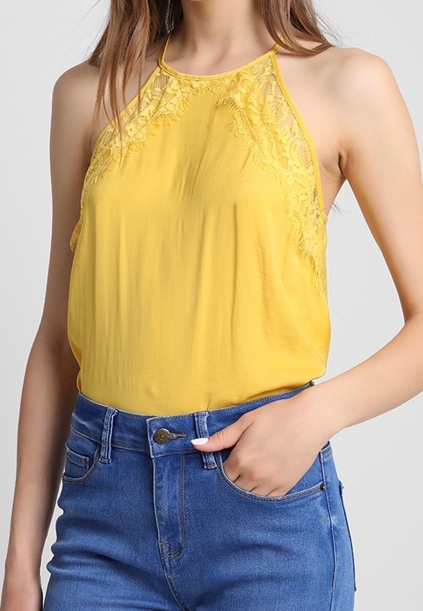 Femme portant un top jaune sans manches à col halter avec des détails en dentelle, rentré dans un jean bleu taille haute, main posée dans la poche avant.