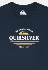 Sötétkék pamut póló, nagy fehér és arany Quiksilver logóval és felirattal, kerek nyakkal és puha tapintással.