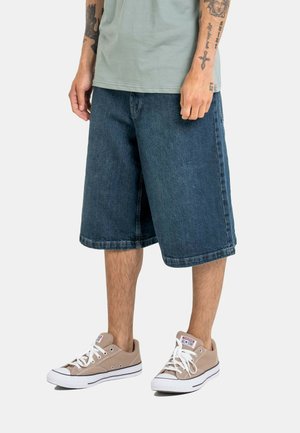 Man wearing loose dark denim shorts, light gray T-shirt, and beige Converse sneakers with visible arm tattoos.