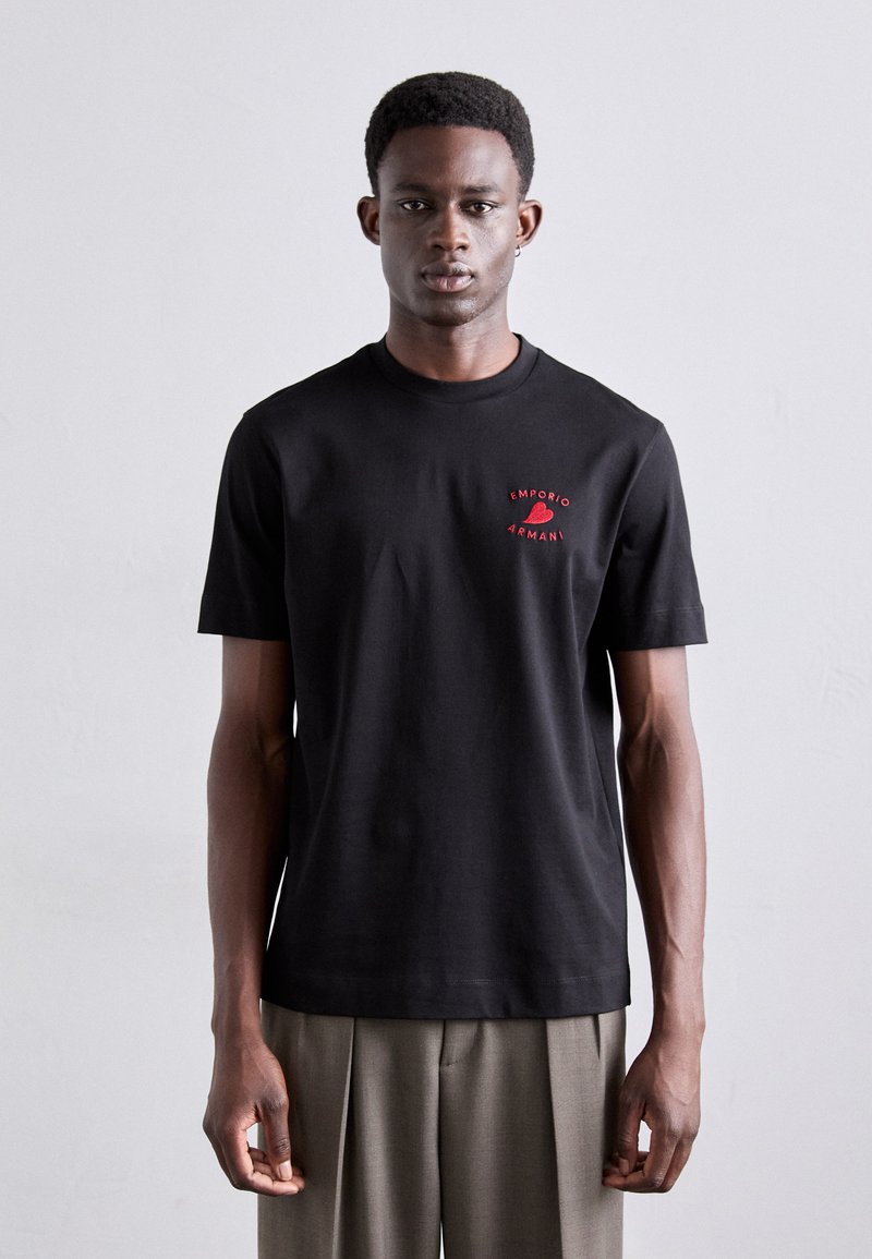 T-shirt en coton noir avec col rond et manches courtes, portant un logo rouge brodé sur la poitrine gauche. Le modèle porte un pantalon de couleur claire.