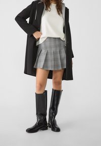 Cappotto nero su una maglietta bianca, gonna svasata a quadri grigi, stivali neri lucidi al ginocchio con tacco spesso, che mettono in mostra un outfit moderno.