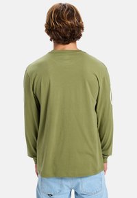 Langärmliges, olivgrünes Hemd aus weichem Baumwollstoff mit lockerem Schnitt. Verfügt über einen Rundhalsausschnitt und sichtbare Nähte entlang der Kanten.