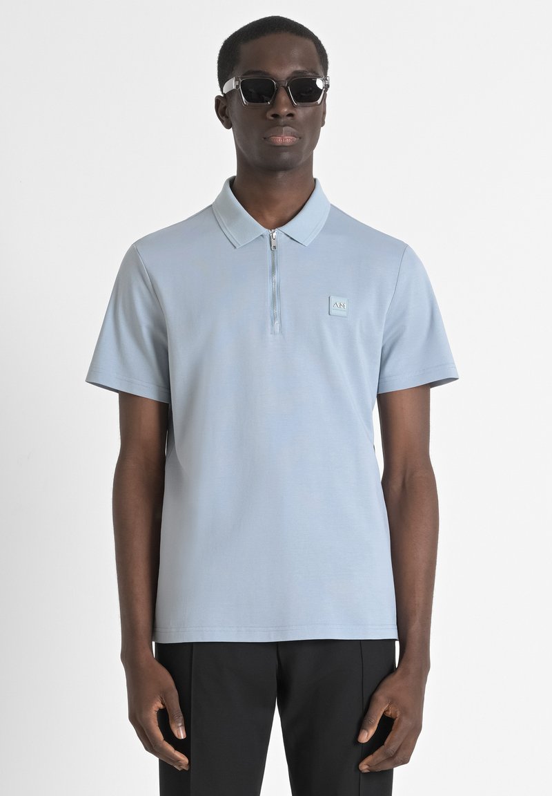 Lichtblauw poloshirt met een halve rits, korte mouwen en een kleine logopatch op de borst, gedragen met donkere broek.