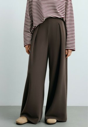 Personne portant un pantalon marron foncé taille haute à jambes larges, des chaussures beiges et un haut rayé bordeaux et blanc à manches longues avec les manches baissées.