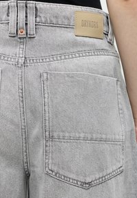 Gros plan sur une personne portant un jean gris clair avec une poche arrière et une étiquette de marque Drykorn couleur beige sur la ceinture.