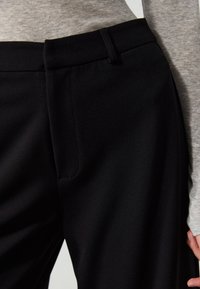 Pantalon noir sur mesure avec une texture lisse et un design épuré. Il présente un devant plat avec des poches et une ceinture structurée.