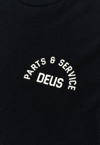 Black cotton t-shirt featuring white curved text: "PARTS & SERVICE" above "DEUS." Round neckline and soft texture.