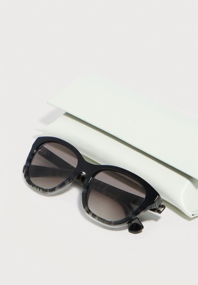 Lunettes de soleil noires oversize avec verres dégradés reposant devant un étui rectangulaire blanc fermé, sur une surface blanche.