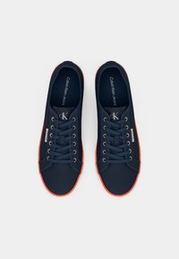 Navy fabric sneakers with red accents, round toe, and white rubber sole. Features laces and "Calvin Klein Jeans" branding on tongue.