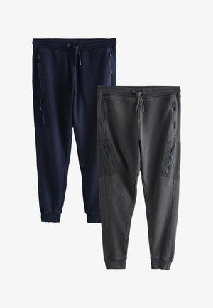 Bărbat purtând pantaloni sport bleumarin cu buzunare laterale și adidași albi Nike, stând în fața unui fundal alb.