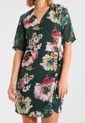 Vrouw draagt een donkergroene wikkeljurk met bloemen in roze, geel en wit en korte mouwen, staand met armen ontspannen langs het lichaam.