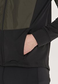 Svart träningsjacka med en texturerad olivgrön panel högst upp. Har en ficka med dragkedja på sidan och åtsittande muddar.