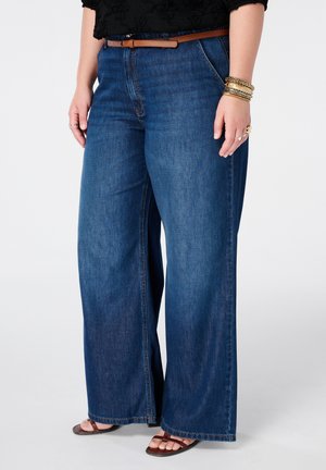 Personne portant un jean bleu à jambes larges avec une ceinture marron, un haut noir, plusieurs bracelets, des bagues et des sandales ouvertes marron sur fond blanc.