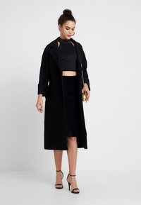 Langer schwarzer Wollmantel mit Spitzkragen, offener Vorderseite und zwei Taschen. Das Modell trägt ein kurzes schwarzes Oberteil und einen Cordrock, kombiniert mit schwarzen High Heels.