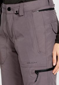Pantalones impermeables de color púrpura claro con un corte ajustado, que cuentan con bolsillos con cremallera, cierre de botón y detalles de costura. Logo de la marca visible.