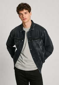 Crna traper jakna klasičnog kroja, s dva džepa na prsima, kopčanjem na dugmad i blago iznošenim detaljima, uparena s svijetlosivom majicom.