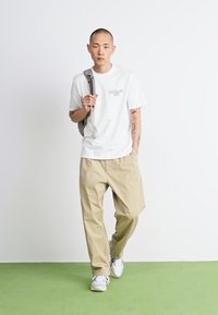 White short-sleeve t-shirt, beige loose-fitting pants, gray sneakers, black backpack with shoulder strap, standing on green floor against a white wall.