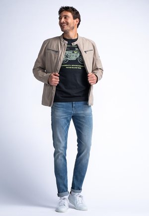 Light beige jacket with zippered pockets, black graphic t-shirt featuring green design, blue jeans, and white sneakers. Simple, modern look.