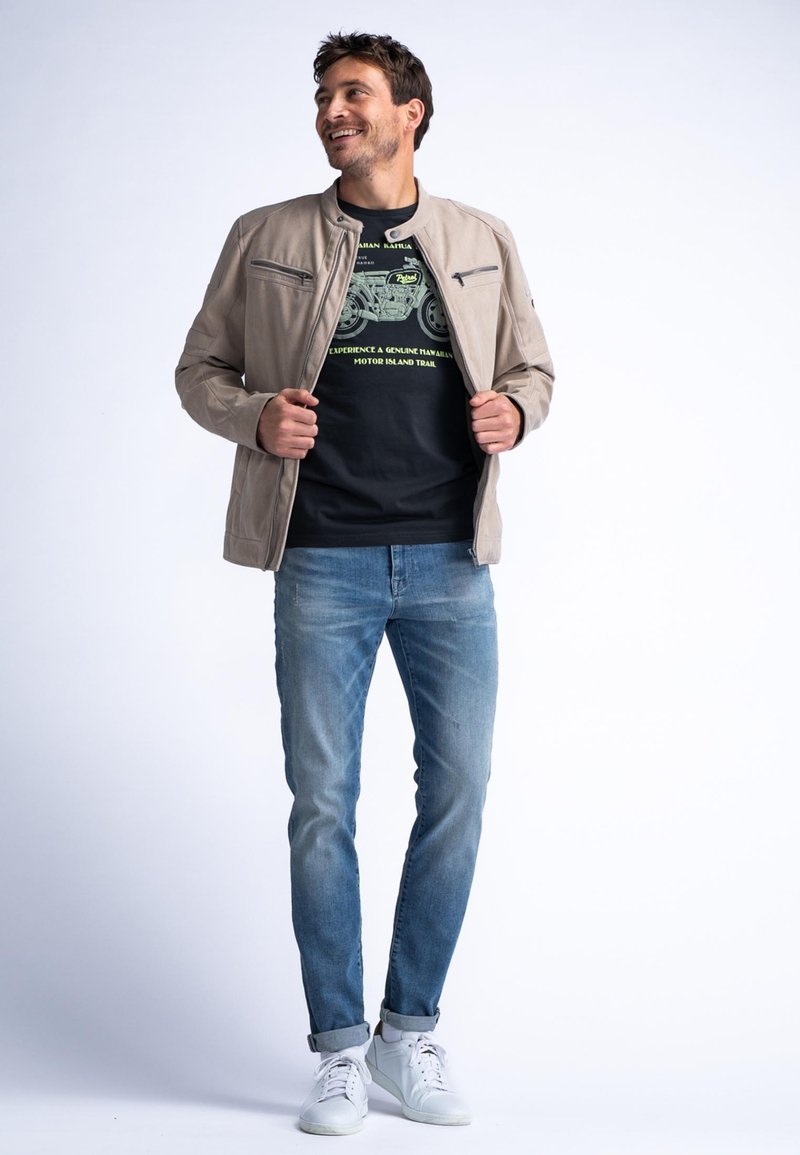 Lichte beige jas met ritszakken, zwart graphic t-shirt met groen ontwerp, blauwe spijkerbroeken en witte sneakers. Eenvoudige, moderne uitstraling.