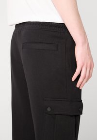 Schwarze Sweatpants aus Baumwolle, mit elastischem Bund, Seitentaschen und einer Klapptasche mit Druckknöpfen am Oberschenkel.
