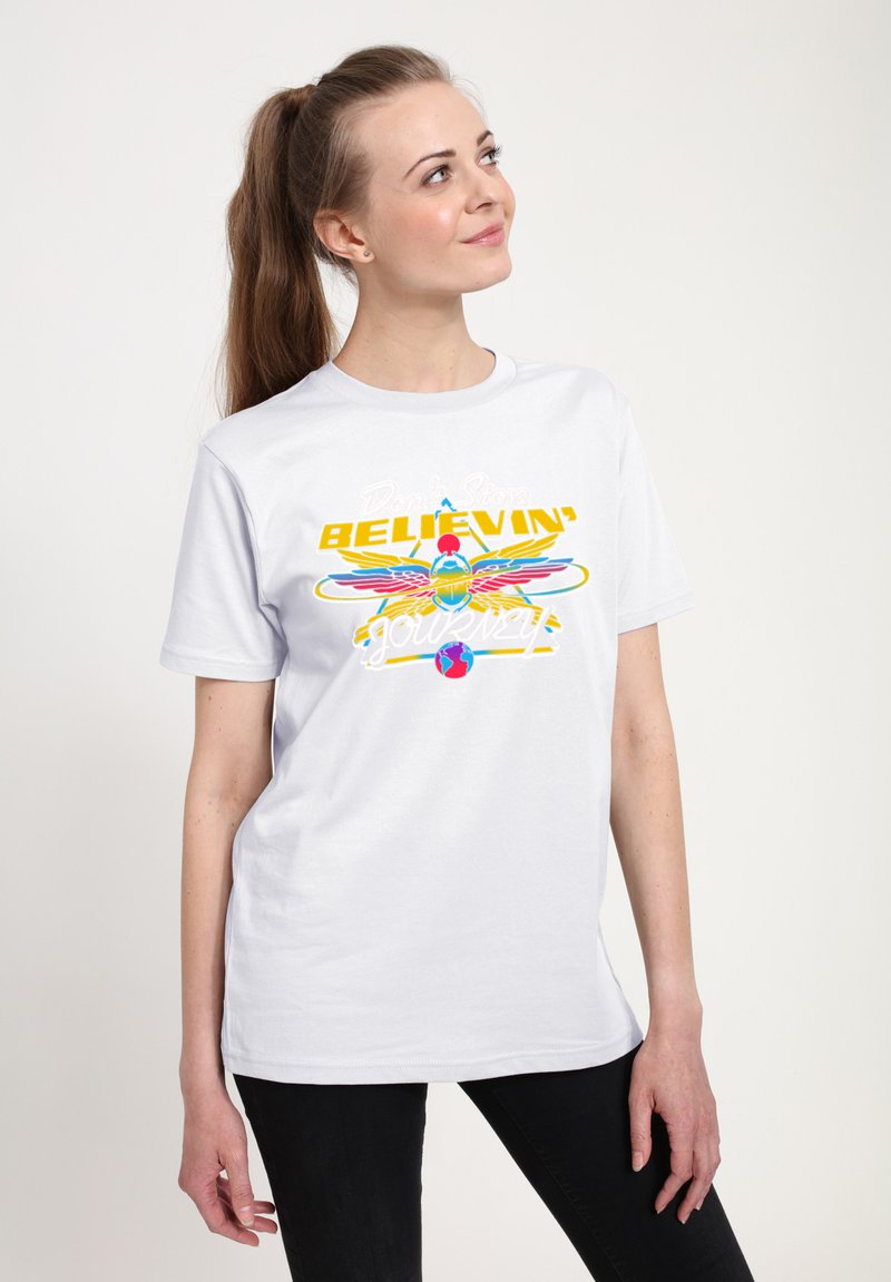 Mujer con coleta que lleva una camiseta blanca con un escarabajo alado colorido y el texto "Don't Stop Believin' Journey", mirando hacia arriba y sonriendo.