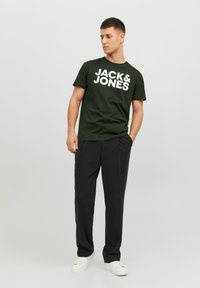 Camiseta de algodón verde oscuro con el texto en blanco "JACK & JONES" en negrita, combinada con pantalones negros y zapatillas blancas, de pie sobre un fondo neutro.