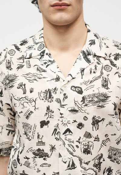 Κοντινό πλάνο ενός ατόμου που φοράει λευκό κοντομάνικο πουκάμισο με μαύρα vintage σχέδια και κείμενο εμπνευσμένα από την Αίγυπτο τυπωμένα παντού.