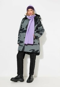 Parka de camuflaje gris con capucha, jeans negros con costura lateral blanca, bufanda morada de canalé y un gorro a juego. Botas de plataforma negra con cordones.