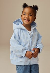 Lys blå lynlås hoodie med blomster mønster, oversized pasform, ribbede manchetter og hætte. Parret med denim jeans.