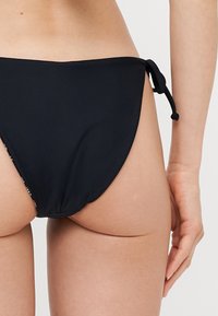 Svart bikini-bunn med et glatt, elastisk stoff. Har en sidekjede lukking og minimal dekning design.