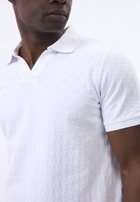 Camisa polo blanca con un patrón texturizado de cuadrados elevados, cuello en V y mangas cortas. La tela parece ligera y transpirable.