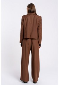 Brązowa marynarka w kroju szytym na miarę i dopasowane spodnie o szerokich nogawkach, wykonane z gładkiego materiału. Marynarka ma klapy i wyrazistą strukturę.