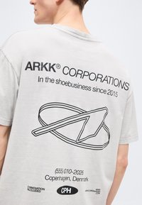 Άτομο που φοράει ανοιχτό γκρι μπλουζάκι με μαύρο κείμενο "ARKK CORPORATIONS", σύνθημα, γραφικό περίγραμμα παπουτσιού και στοιχεία επικοινωνίας στην πλάτη.