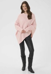 Šviesiai rožinė didelė pončo stiliaus megztinis su įstrižine tekstūra, ilgomis rankovėmis, dėvimas ant juodų odinių aptemptų kelnių ir juodų cowboy stiliaus aulinukų.