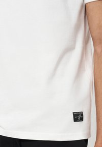Chemise blanche à manches courtes avec une petite étiquette rectangulaire noire près de l'ourlet inférieur présentant un logo d'oiseau et du texte.