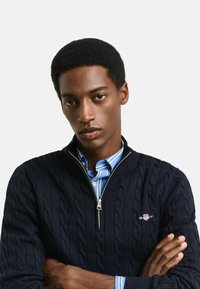 Pull en maille bleu marine avec motif en câble, col zippé, porté par-dessus une chemise rayée bleu clair et blanche. Accessoire : patch logo sur la poitrine.
