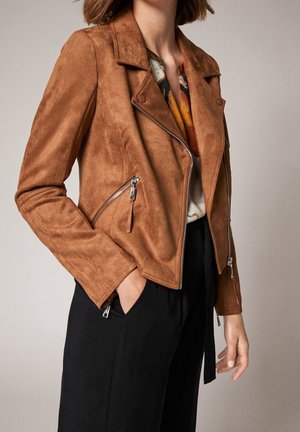 Kvinde iført en brun ruskinds bikerjakke med sølvfarvede lynlåse over en mønstret bluse og sorte bukser, hånd i lommen, foran en ensfarvet baggrund.