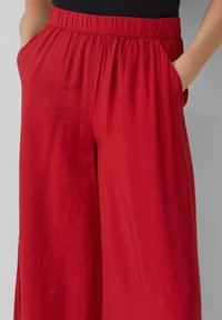 Pantalons larges rouges en tissu texturé, dotés d'une taille élastique et de poches sur les côtés. Fini lisse avec un léger éclat.